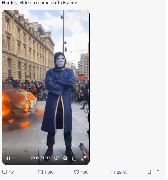 Figure in a long blue coat and mask standing in front of a burning car at a protest